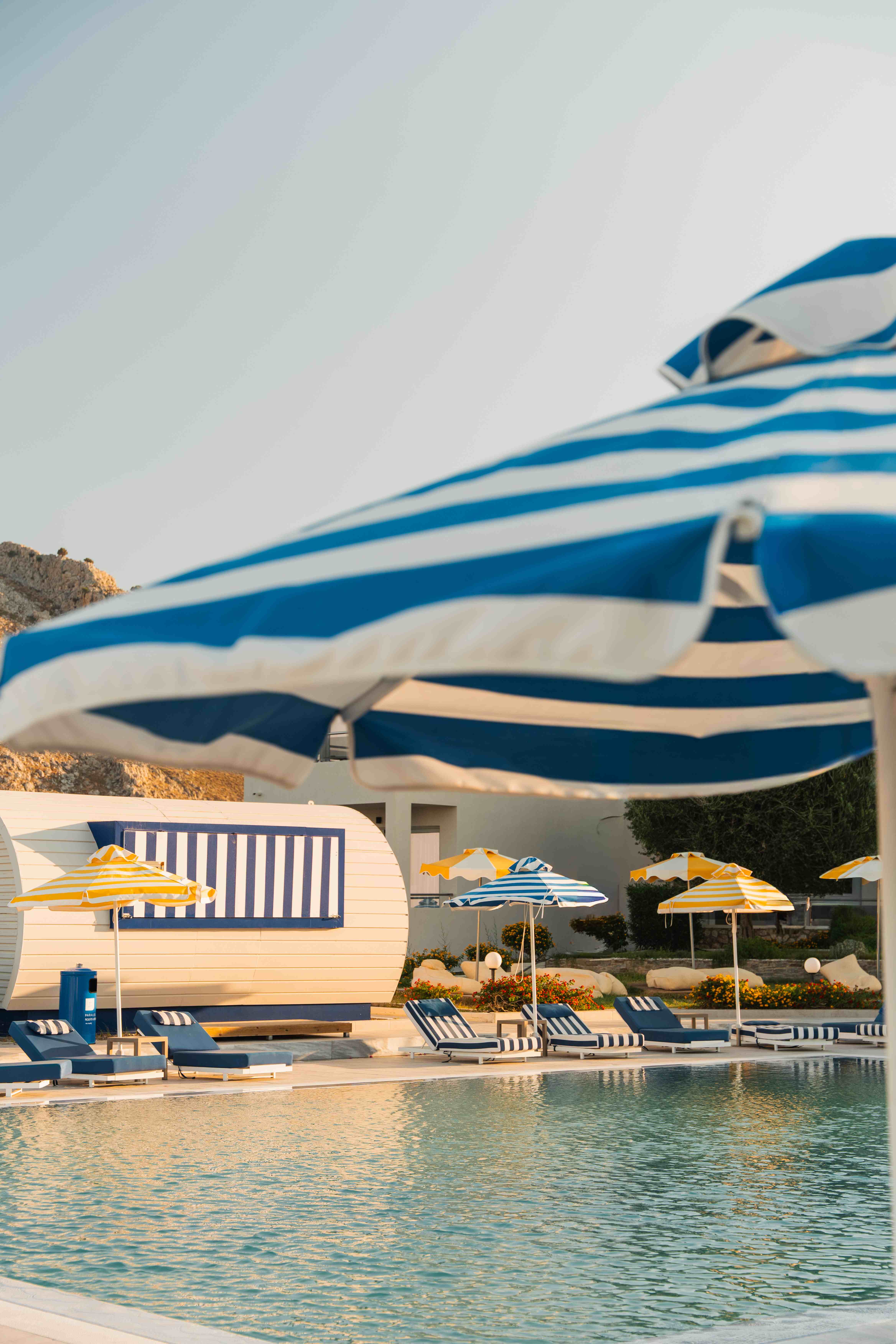 Blue and white striped umbrella in close-up, with the pool, sun loungers, yellow umbrellas and retro caravan bar behind