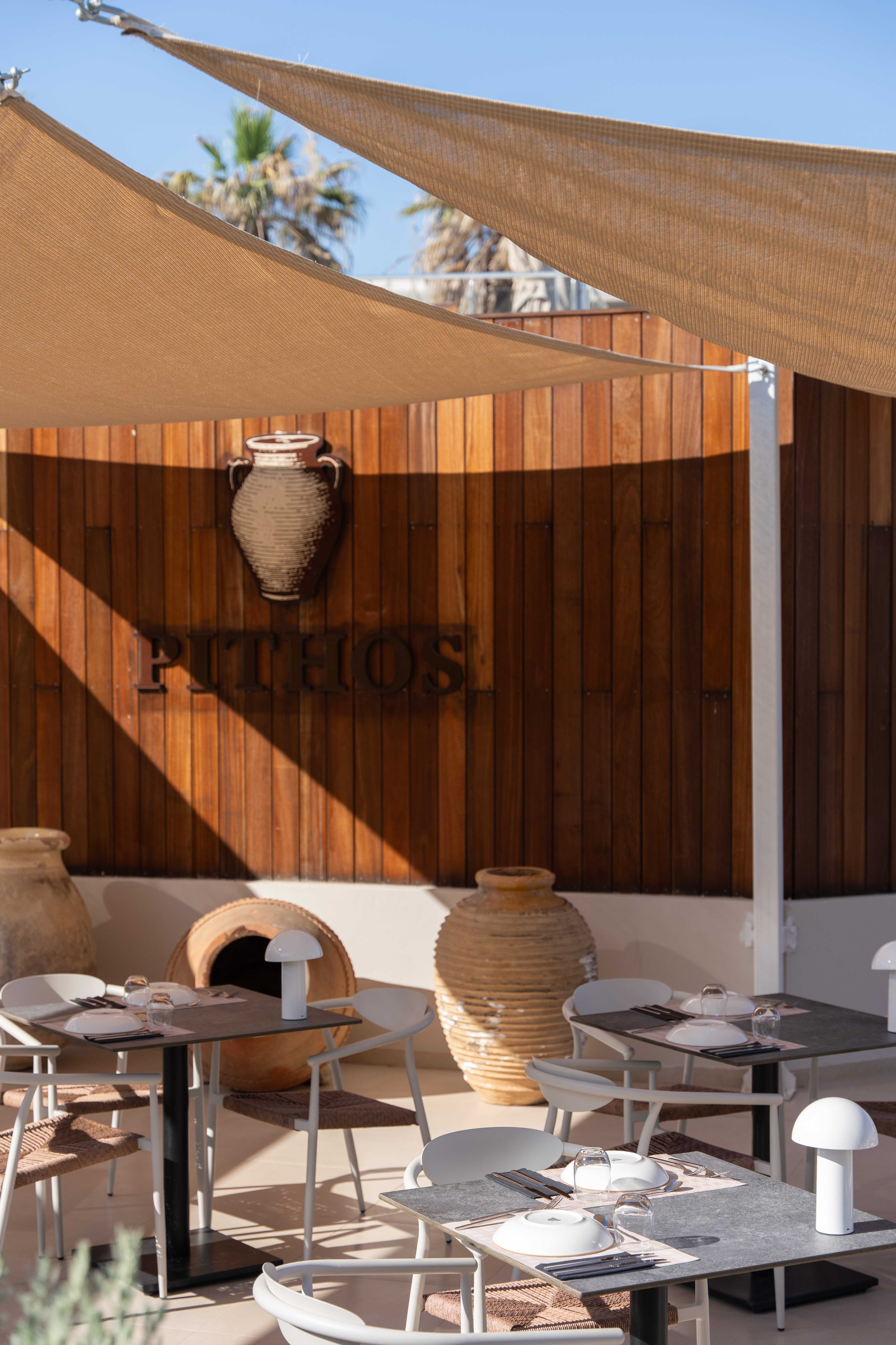 Sunlight and shade on the Pithos wooden wall with a hanging clay jar, terracotta pots and set tables under sail shades