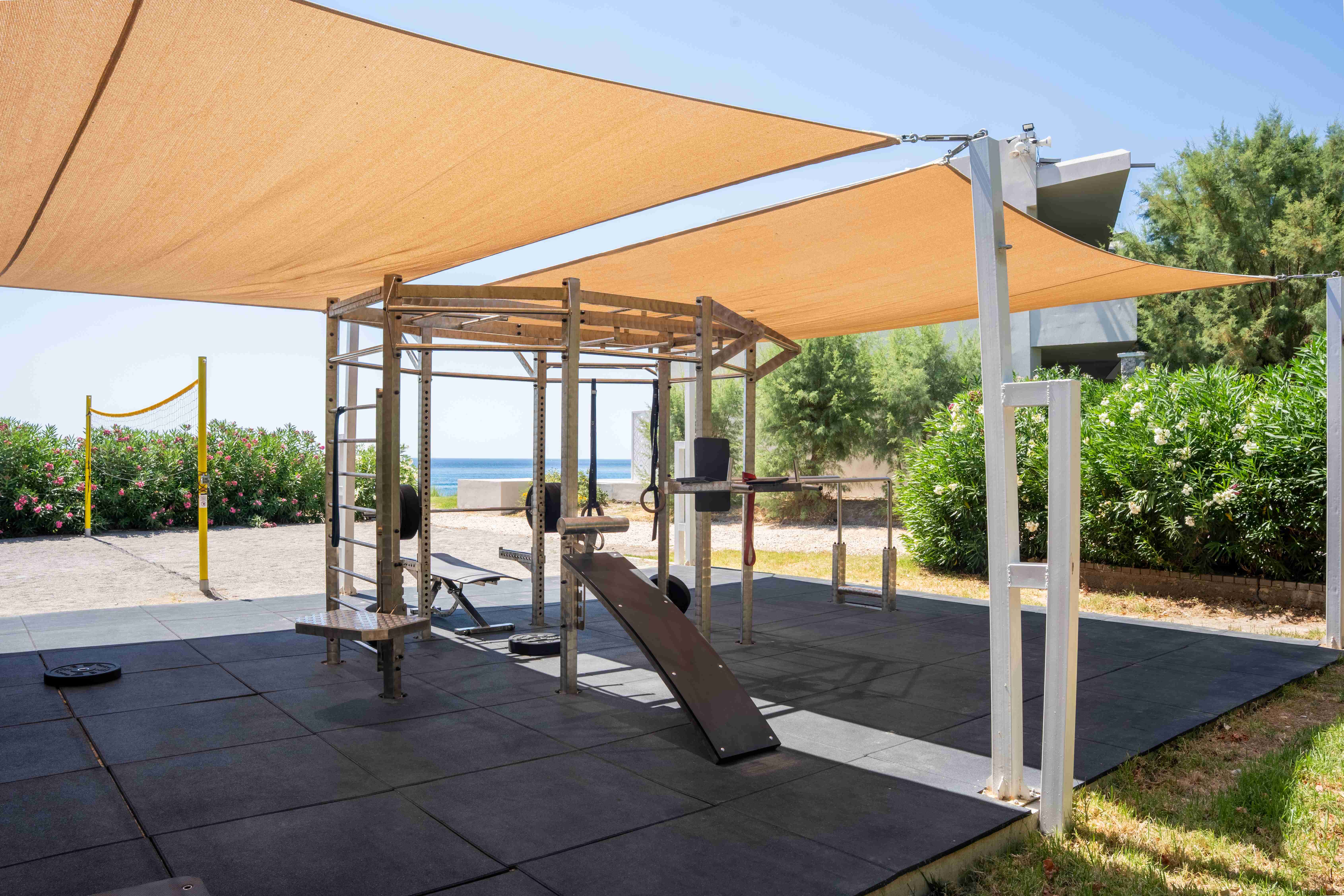 Outdoor fitness area under sail shades, with a training rig, incline bench and rubber flooring among green shrubs