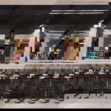 Modern hotel lobby bar with high stools, backlit shelves of liquor bottles and dark tiled counter under black ceiling panel