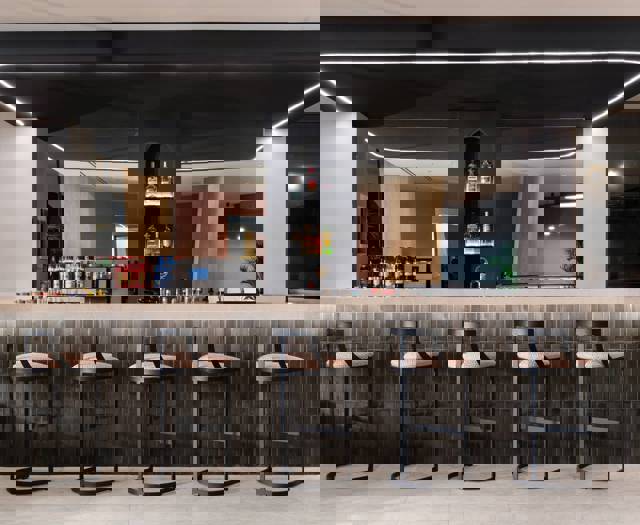 Modern hotel lobby bar with high stools, backlit shelves of liquor bottles and dark tiled counter under black ceiling panel