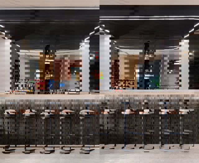 Modern hotel lobby bar with high stools, backlit shelves of liquor bottles and dark tiled counter under black ceiling panel