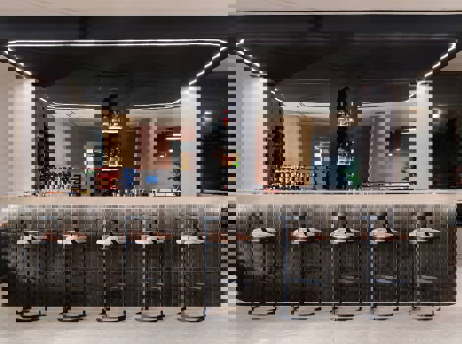 Modern hotel lobby bar with high stools, backlit shelves of liquor bottles and dark tiled counter under black ceiling panel