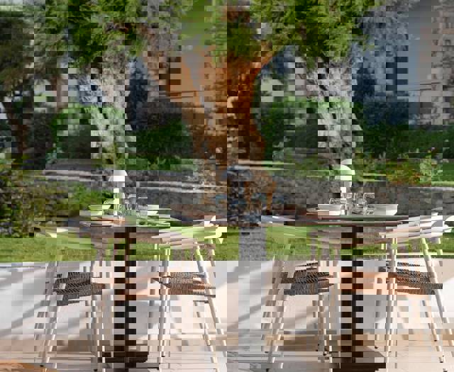 Outdoor dining table with four white chairs set near a garden with a large tree