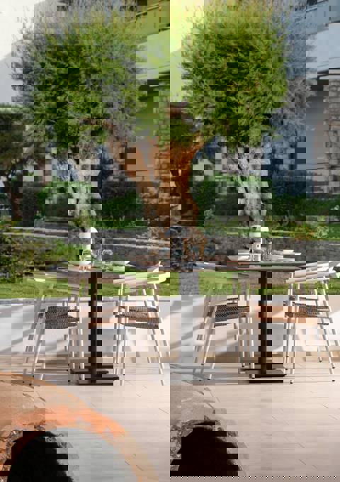Outdoor dining table with four white chairs set near a garden with a large tree