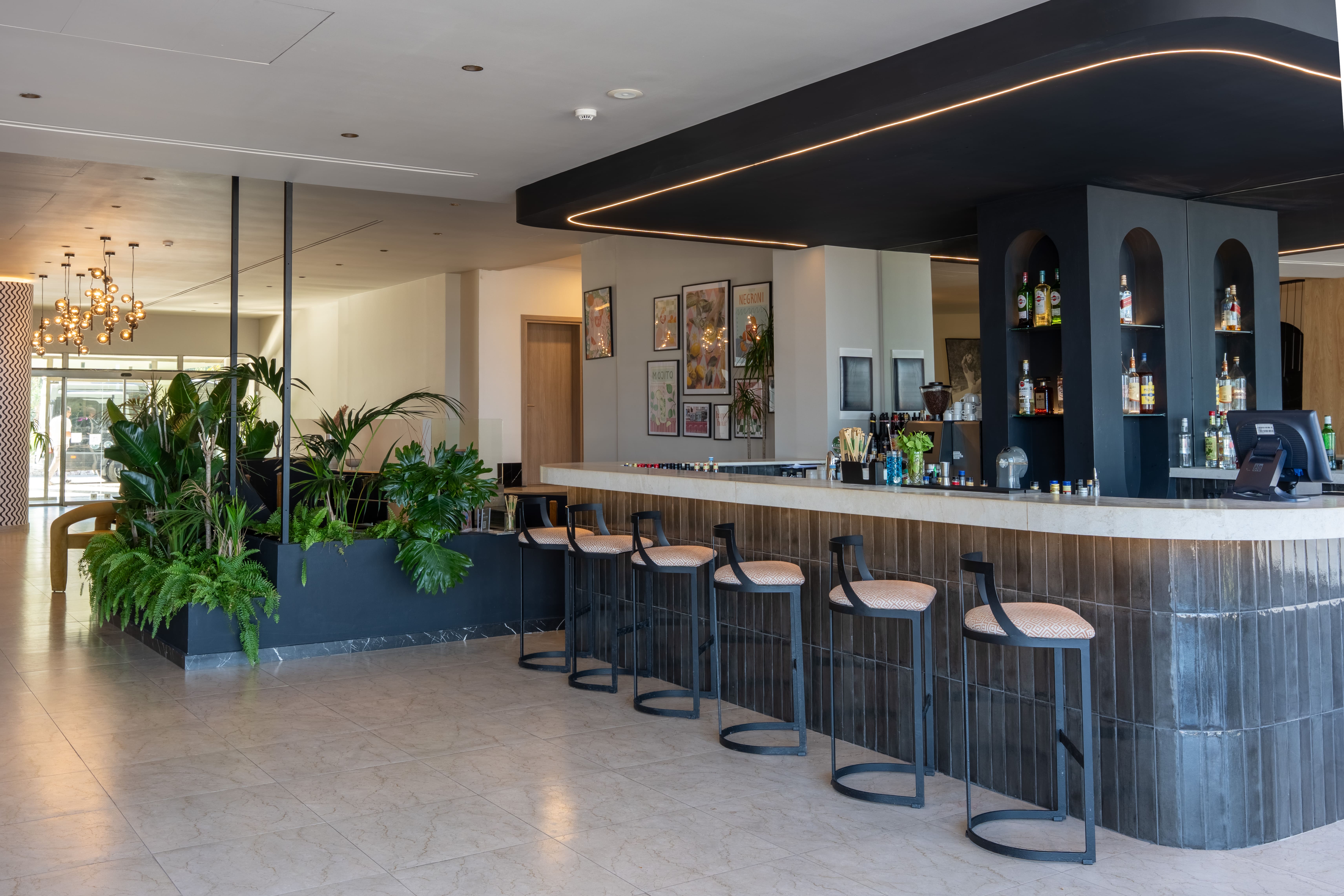 Bar with glossy dark tiles, black stools and a neon-edged canopy over arched shelves, beside lush plants and framed art