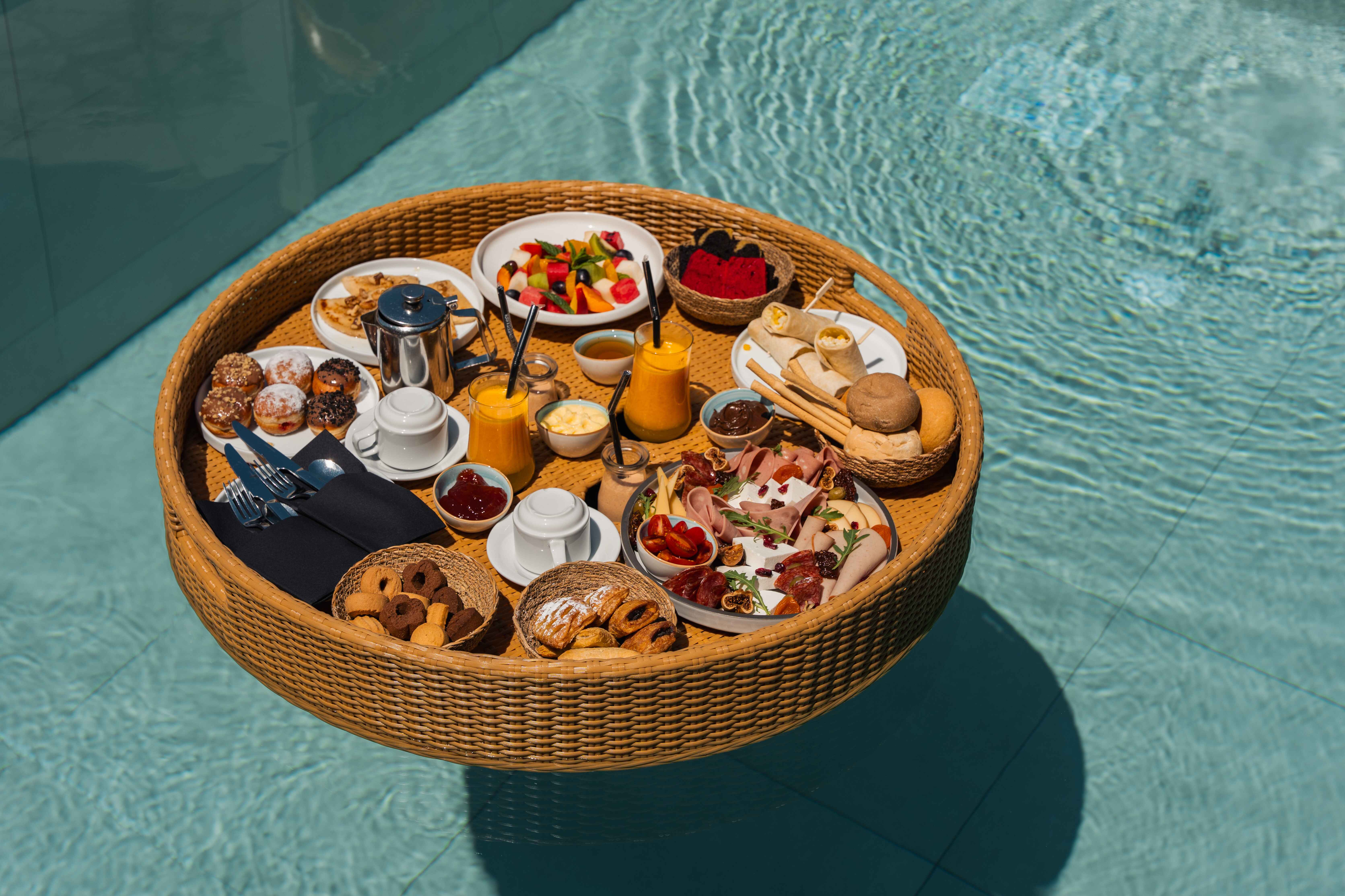 Wicker floating breakfast tray on the pool with fruit, cold cuts, pastries, cookies, orange juice and a pot of coffee