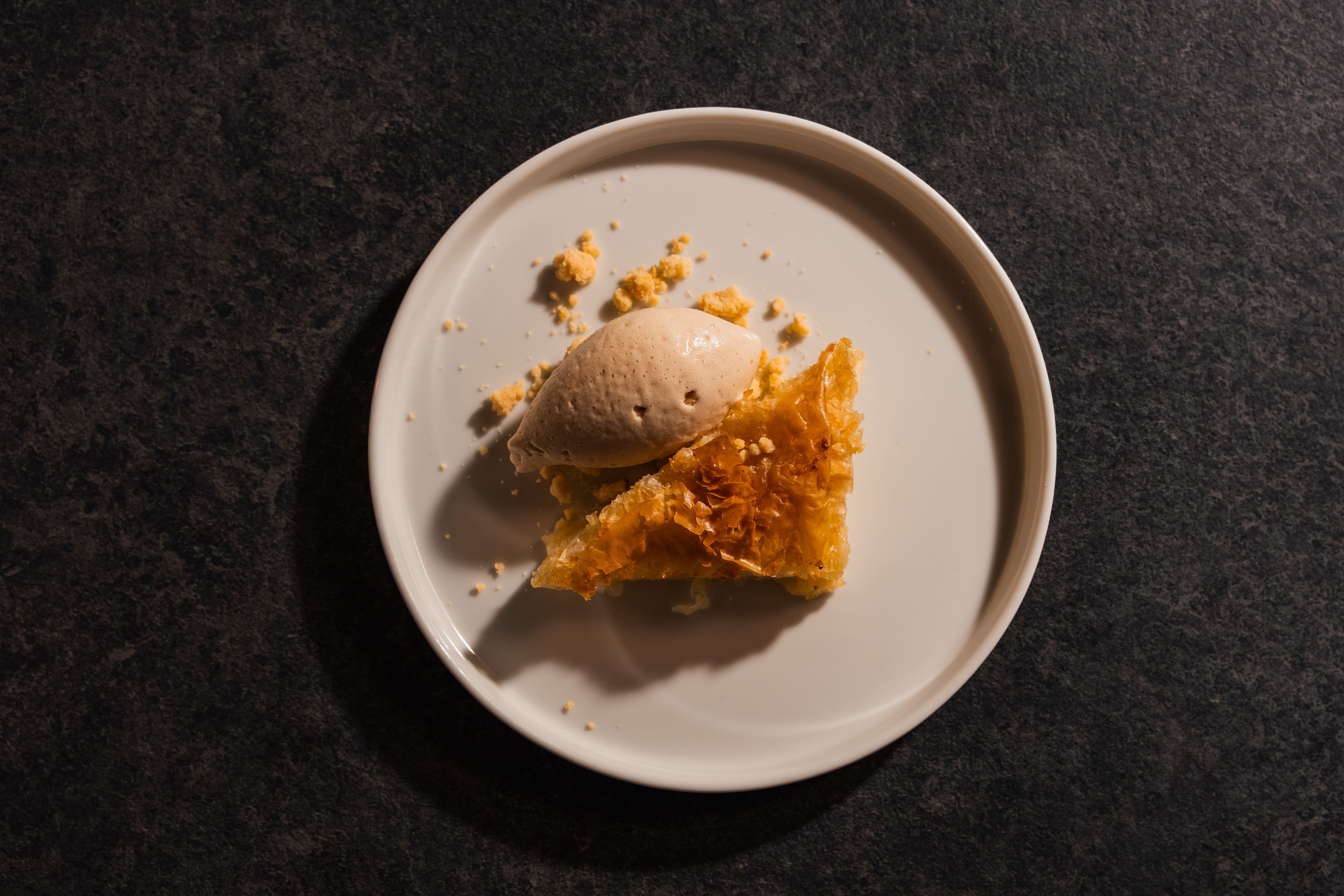 Quenelle of caramel ice cream on crisp kataifi pastry with golden crumble, served on a white plate over dark stone