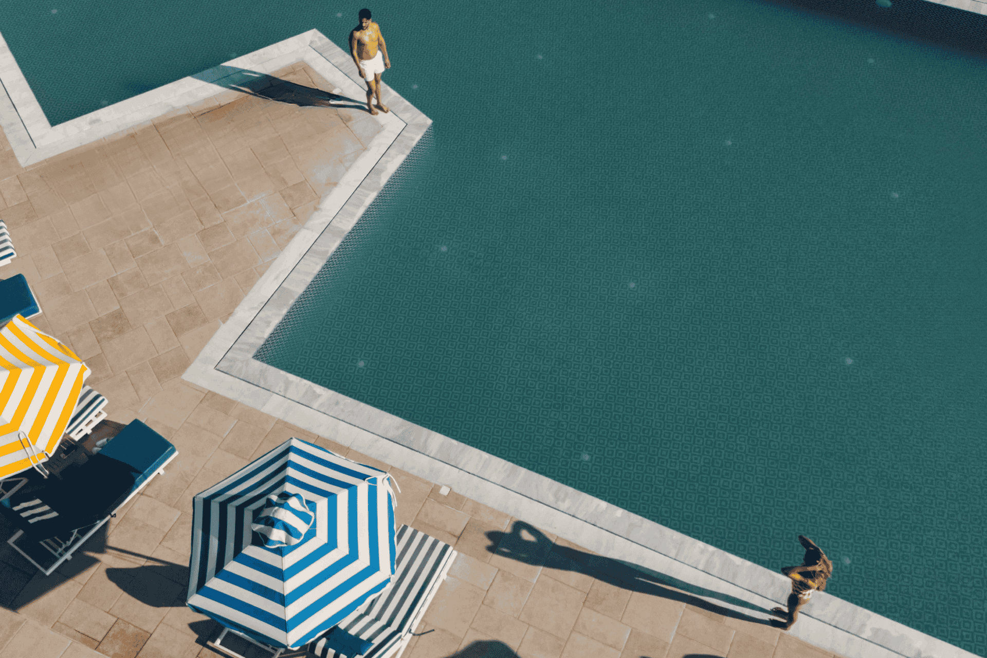 Overhead view of the angular pool's deep teal water, two people casting long shadows at its edges by striped umbrellas