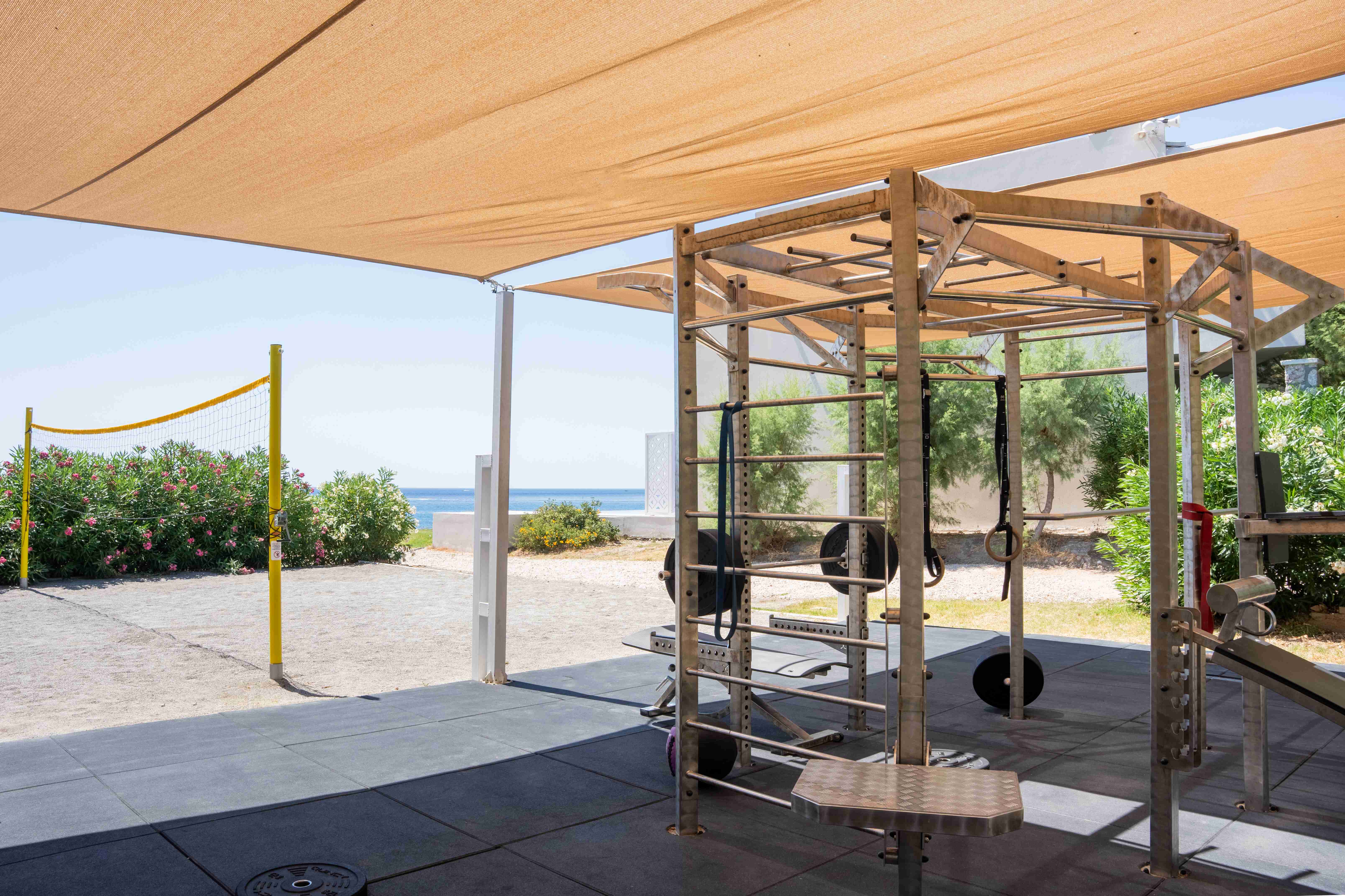Shaded outdoor gym with a training rig, weights and suspension straps beside a beach volleyball court and the sea