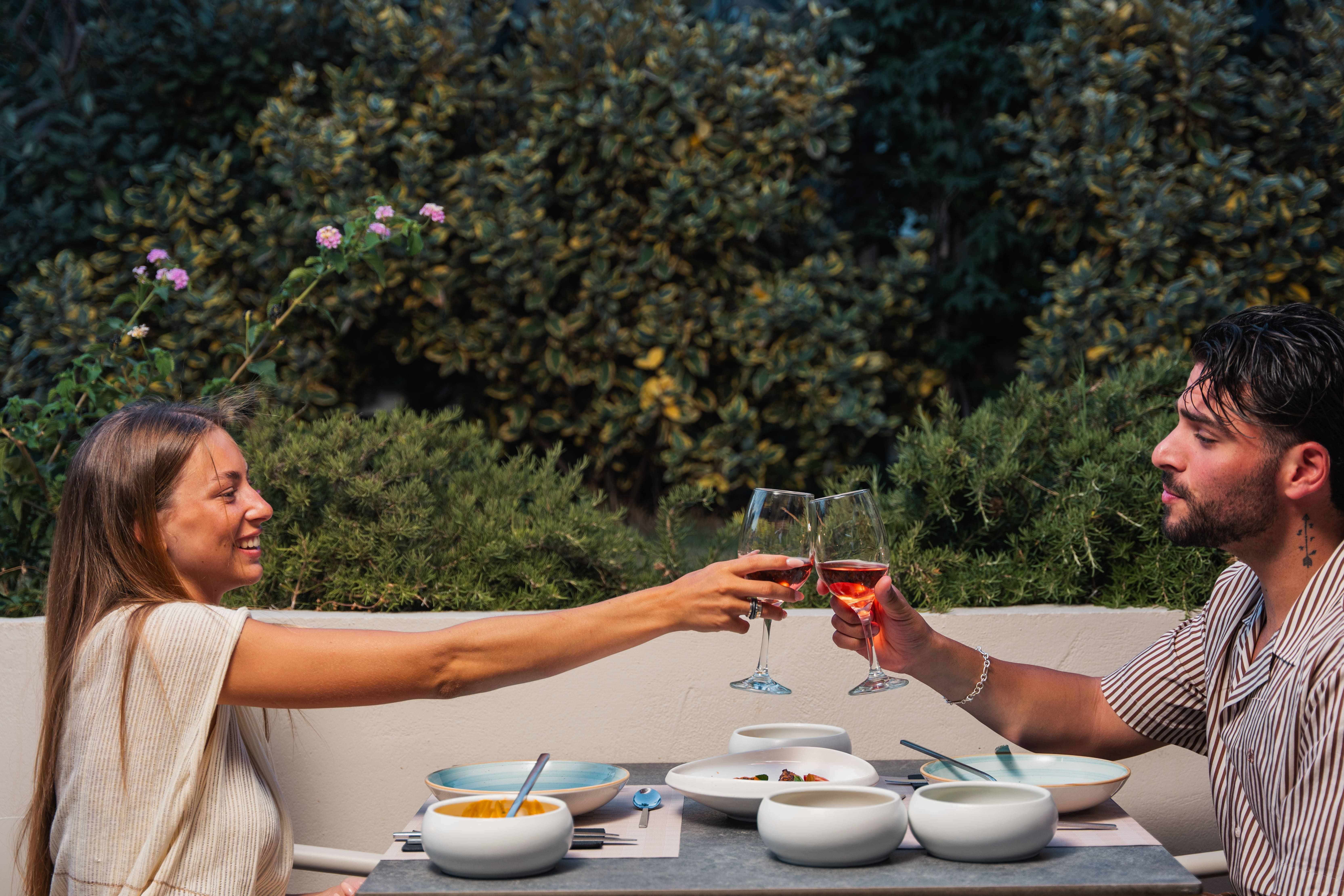 Smiling couple toasting with glasses of rosé over a table of shared dishes, against a lush green hedge with pink flowers